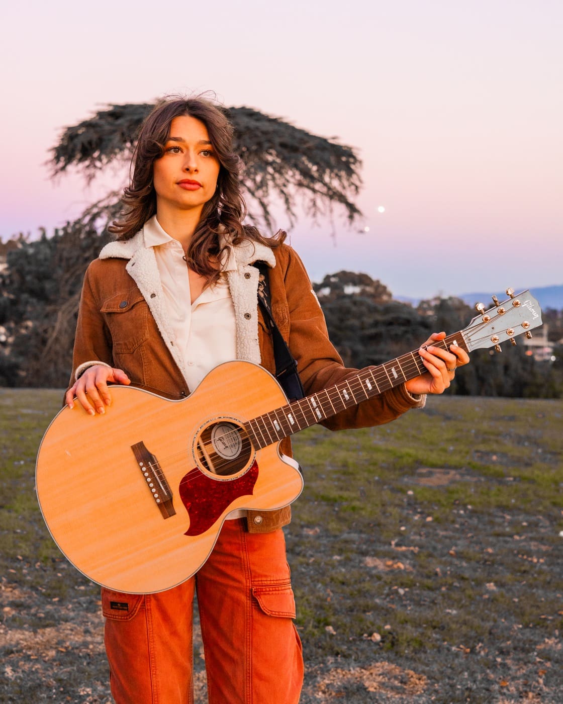 Adana holding an acoustic guitar outdoors at sunset
